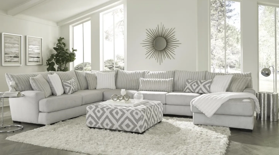 Modern living room with a large gray L-shaped sofa, geometric-patterned ottoman, fluffy rug, and decorative pillows. A sunburst mirror and several paintings adorn the white walls.